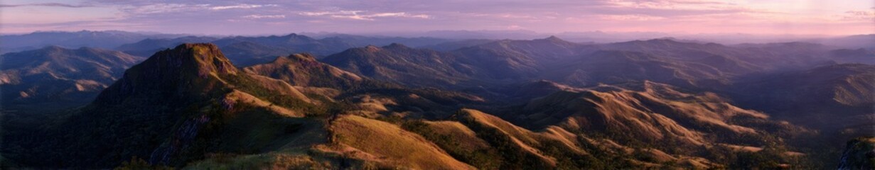 Fototapeta premium Panoramic view of a mountain range at sunset, with the foreground showing a closer, detailed view of a peak, while the background displays a hazy expanse of mountains under a soft, purplish sky