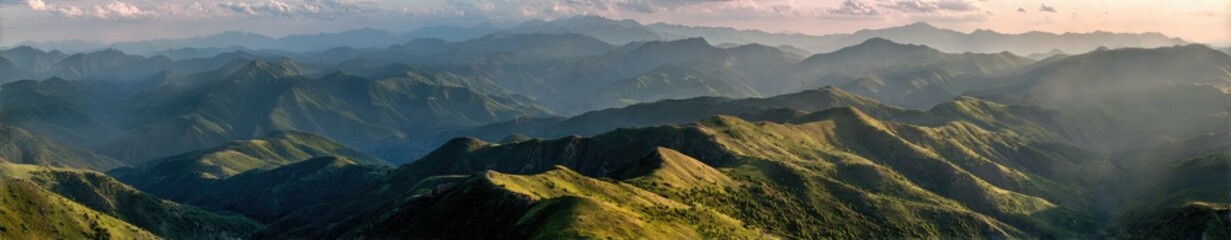 Fototapeta premium Panoramic view of a vast mountain range, bathed in the soft light of dawn or dusk. Rolling hills and valleys stretch into the distance, shrouded in a delicate haze