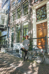 Beautiful elegant woman is enjoying summer vacation in Europe landmark Amsterdam city, while wearing fashion black and white knit dress, red military hat holding red roses, posing in front of bicycle.