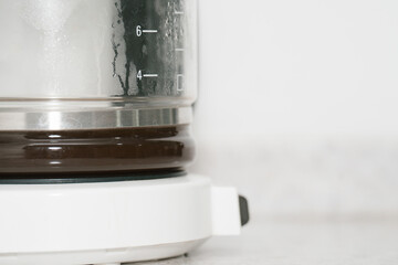 A white drip coffee maker with brewed coffee visible through the glass carafe. The steam condensation and measurement markings emphasize the brewing process. White background with ample copy space