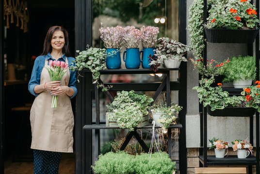 Woman florist reopening shop after covid-19 pandemic, delivery service for clients. Happy young female in apron holds bouquet of tulips near front door of store with vases and pots of diverse flowers