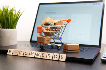 Miniature shopping cart full of cardboard boxes is standing on laptop keyboard with e-commerce...