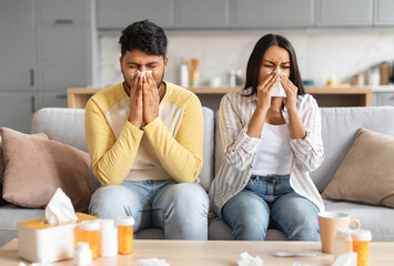 Sick indian couple sitting on sofa, blowing noses and suffering from colds, ill eastern spouses...