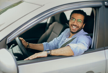 Cheerful long-haired arab guy with glasses driving right wheel auto, looking through open window...