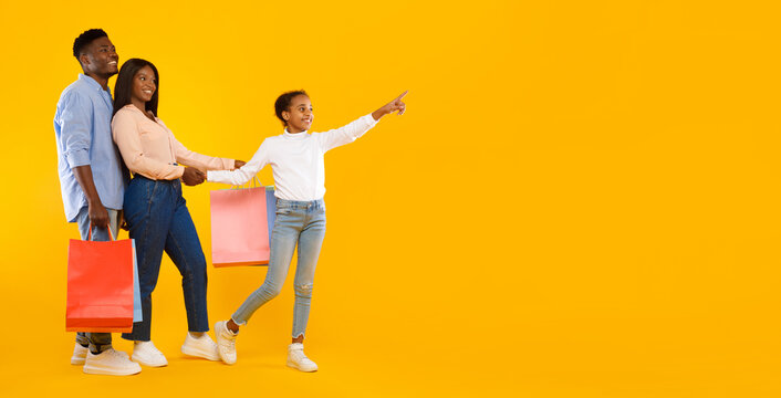 Family Shopping Concept. Full length body size portrait of African American cheerful people holding shopping bags with new clothes, excited little girl pointing aside at copy space, yellow studio wall