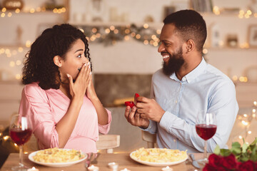 Proposal Date Concept. Smiling black boyfriend holding open box, showing engagement ring to excited...
