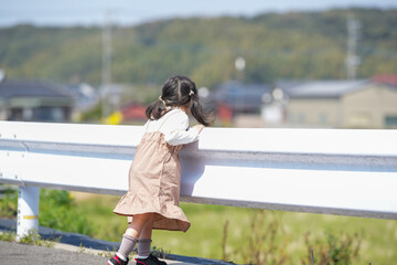 A 3-year-old Japanese girl stands beside a white guardrail, quietly gazing at the countryside....