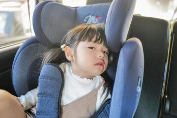 A 6-month-old baby and a 3-year-old Japanese girl sit in car seats in the back seat, gazing forward...