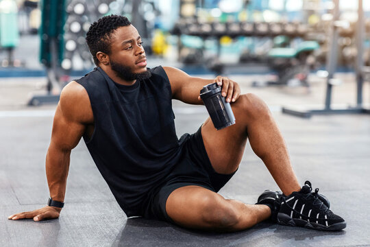Handsome african american bodybuilder holding bottle with water or sports drink, sitting on floor at gym, resting, empty space. Smiling black muscular man having break while exercising at modern gym