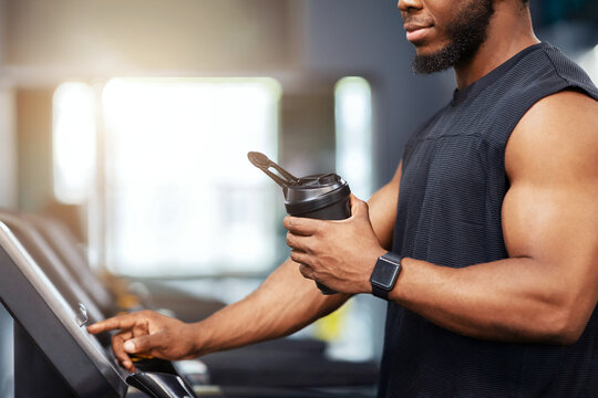 Unrecognizable black guy sportsman with protein drink standing on treadmill, side view, copy space. Cropped of muscular african american man having jogging work out, drinking water, sun flare