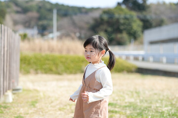 A cheerful 3-year-old Japanese girl with twin-tails is spending a sunny spring afternoon in March, feeding green grass to a friendly goat in a quiet countryside field bathed in soft warm sunlight.
