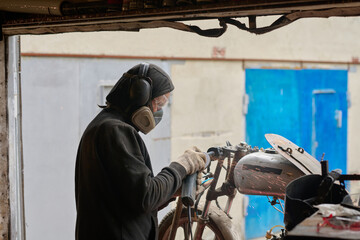Middle aged Caucasian man wearing protective mask and headphones repairing custom motorcycle in...