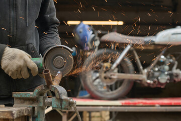 Caucasian young adult man using angle grinder to cut metal pipe in workshop, sparks flying while custom motorcycle stands in background, hands wearing protective gloves, focused on repair work