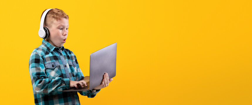 Excited redhead boy with wireless headset playing on laptop, yellow studio background. Amazed kid in casual outfit using modern powerful gadget for video games, playing online, copy space - Powered by Adobe