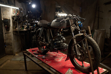Custom motorcycle on workbench in dimly lit workshop surrounded by tools and mechanical parts,...