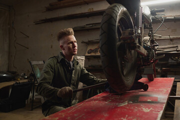 Caucasian young adult man repairing custom motorcycle in workshop using tools, focusing on rear wheel assembly, working alone in garage surrounded by equipment and bike parts