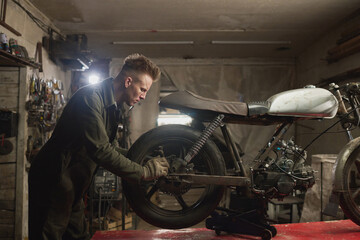 Young adult Caucasian man repairing custom motorcycle in workshop, focusing on adjusting rear wheel and suspension components, surrounded by tools and mechanical equipment