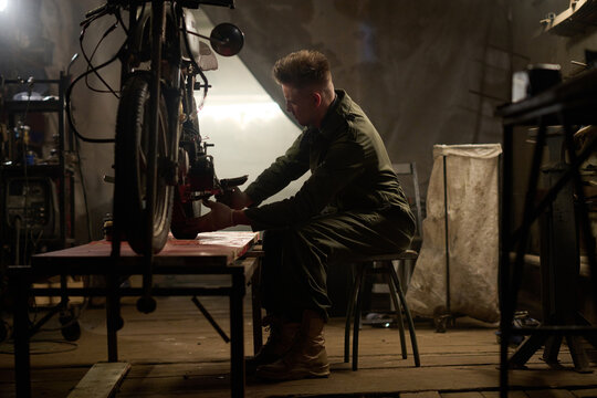 Caucasian young adult man repairing custom motorcycle in workshop, sitting on chair and adjusting bike components, focused on mechanical work in dimly lit garage environment - Powered by Adobe