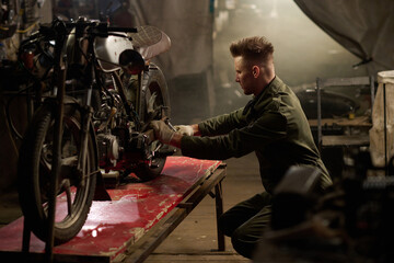 Caucasian young adult man kneeling beside custom motorcycle repairing engine components in workshop...