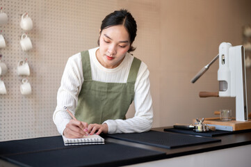 Asian female barista in green apron writing a drink ingredients in notebook at coffee counter bar