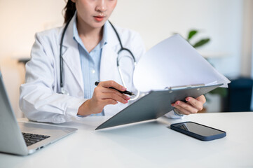 Asian female doctor reading and pointing a pen at clipboard with laptop on table in diagnosis room.