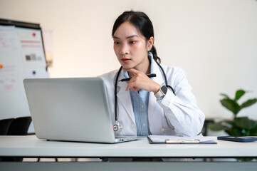 Asian female doctor holding pen under chin and looking closely at laptop on table in diagnosis room.