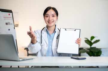 Asian female doctor showing a paper on clipboard explaining with laptop at table in diagnosis room.