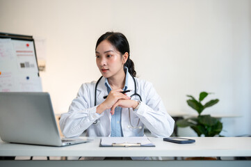 Asian female doctor holding hands and pen under chin looking at laptop on table in diagnosis room.