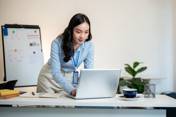 Female office worker or businesswoman standing and leaning over a working desk to look at laptop.