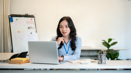 Female office worker businesswoman holding pencil and looking at laptop sitting at working table.