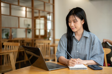 Smiling asian woman holding pen writing in notebook while looking at laptop on cafe's wooden table.