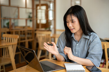 Asian woman talking or explaining and looking at laptop with notebook and phone on cafe wooden table