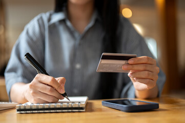 Close up of a woman holding credit or debit card and a pen writing in notebook sitting at cafe table