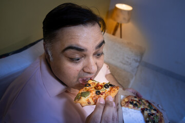 Hungary overweight fat indian man eating cheese pizza at home. Plus size guy enjoy fatty junk food. Obese male taking a bite of high calorie meal. Obesity and weight gain. Midnight craving. Closeup
