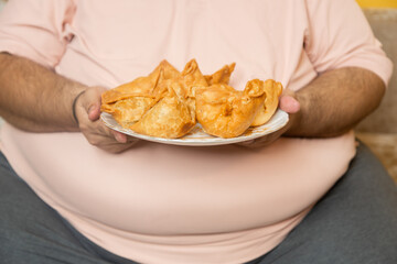 Overweight fat indian man holding plate of unhealthy samosa junk food. Plus size guy eating fried snack. Obese male enjoy high calorie meal. Obesity and weight gain. Closeup.