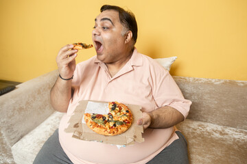 Hungary overweight fat indian man eating cheese pizza. Plus size guy enjoy fatty junk food. Obese male taking a bite of high calorie meal. Obesity and weight gain.