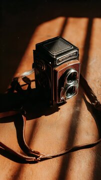 Vintage twin lens reflex camera rests on a tan surface, bathed in warm, dramatic sunlight creating strong shadows. Brown leather strap is partially visible.