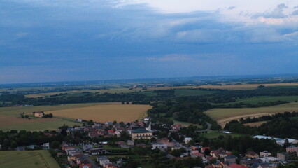Fototapeta premium Aerial drone view of a peaceful rural landscape during summer sunset in Silesia, Czech Republic. The horizon glows with warm colors above rolling fields, small village houses, and distant Jesenik.