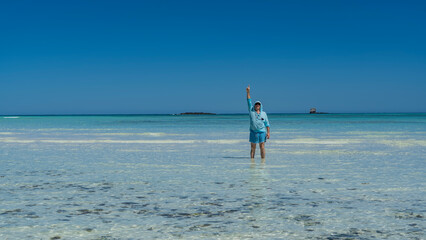 A man in sunglasses and a hooded shirt stands in shallow water in the ocean. The hand is raised up, the finger is pointing into the blue sky. The bottom is visible through the clear aquamarine water. 