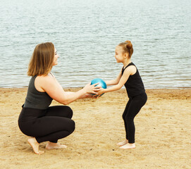 A mother plays ball with her six-year-old daughter on the beach by the lake on a summer day.