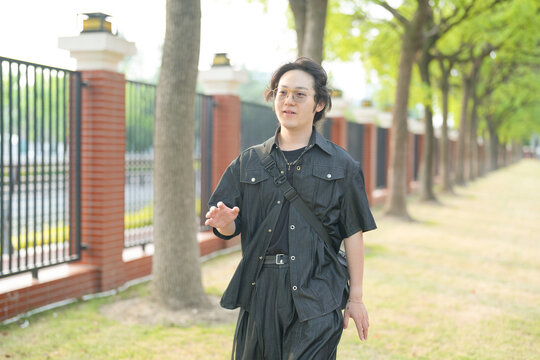 A Chinese man in his 20s, wearing glasses, walks across a university campus on a bright spring day. The calm, peaceful scene is filled with sunshine and fresh greenery in Xuhui District, Shanghai.