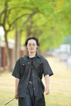 A Chinese man in his 20s, wearing glasses, walks across a university campus on a bright spring day. The calm, peaceful scene is filled with sunshine and fresh greenery in Xuhui District, Shanghai.