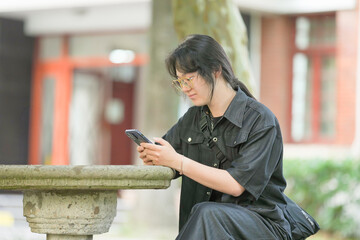 A young Chinese man in his 20s, wearing glasses and a light spring outfit, sits on a wooden bench in the university campus, calmly using his smartphone under the clear blue sky. Xuhui, Shanghai.