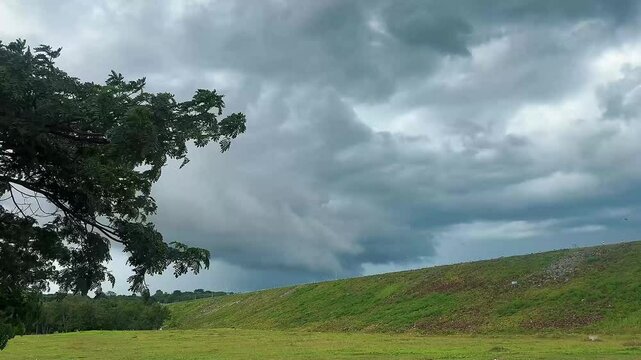 Landscape view of a rural road curving along a dam or reservoir embankment under a dark, dramatic sky filled with gathering storm clouds. A quiet, moody atmosphere before the rain