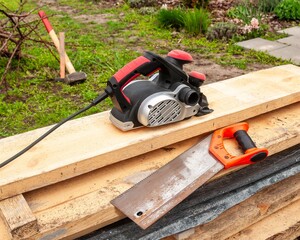 Electric planer and hand saw rest on wooden planks, ready for woodworking outside.