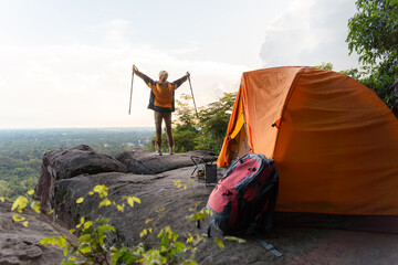 A man is standing on a rock with a backpack and a tent. He is holding two poles