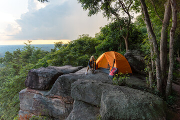 A person is setting up a tent on a rocky hill. The tent is orange and is located near a tree