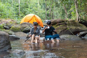Two people sitting in a river, one of them holding a cup. One of them is wearing a plaid shirt
