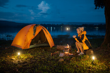 A woman sits by a campfire with a tent in the background. She is wearing a yellow jacket