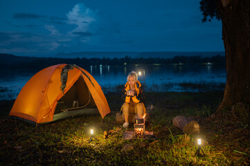 A woman sits by a campfire in the woods. She is wearing a yellow jacket. The fire is lit and there...